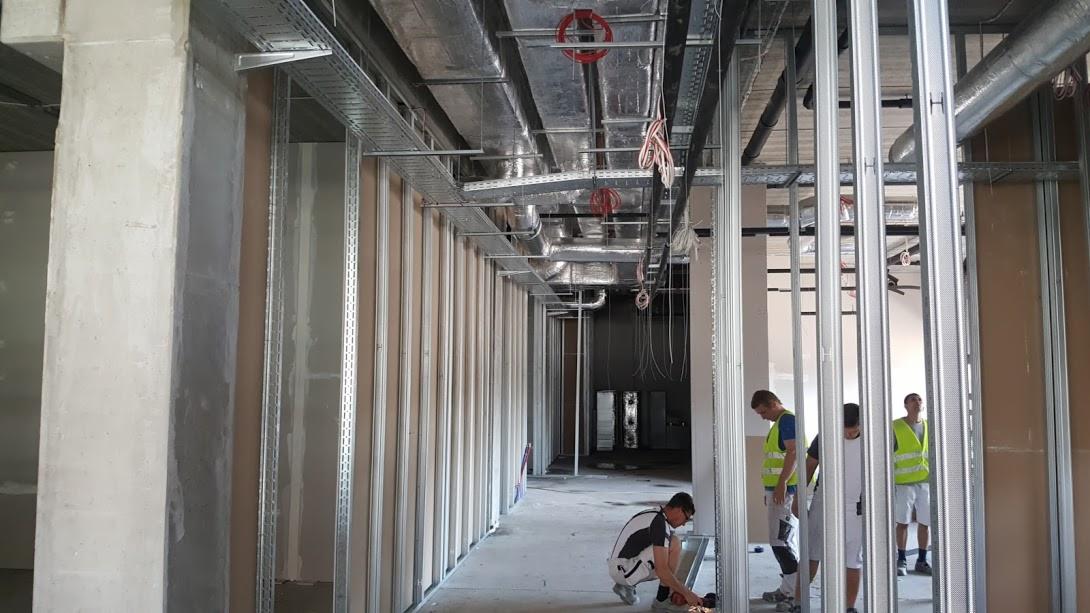 Wnętrze w trakcie prac wykończeniowych: montaż metalowej konstrukcji pod płyty gipsowo-kartonowe, widoczne instalacje pod sufitem i pracujący monter w okularach ochronnych.