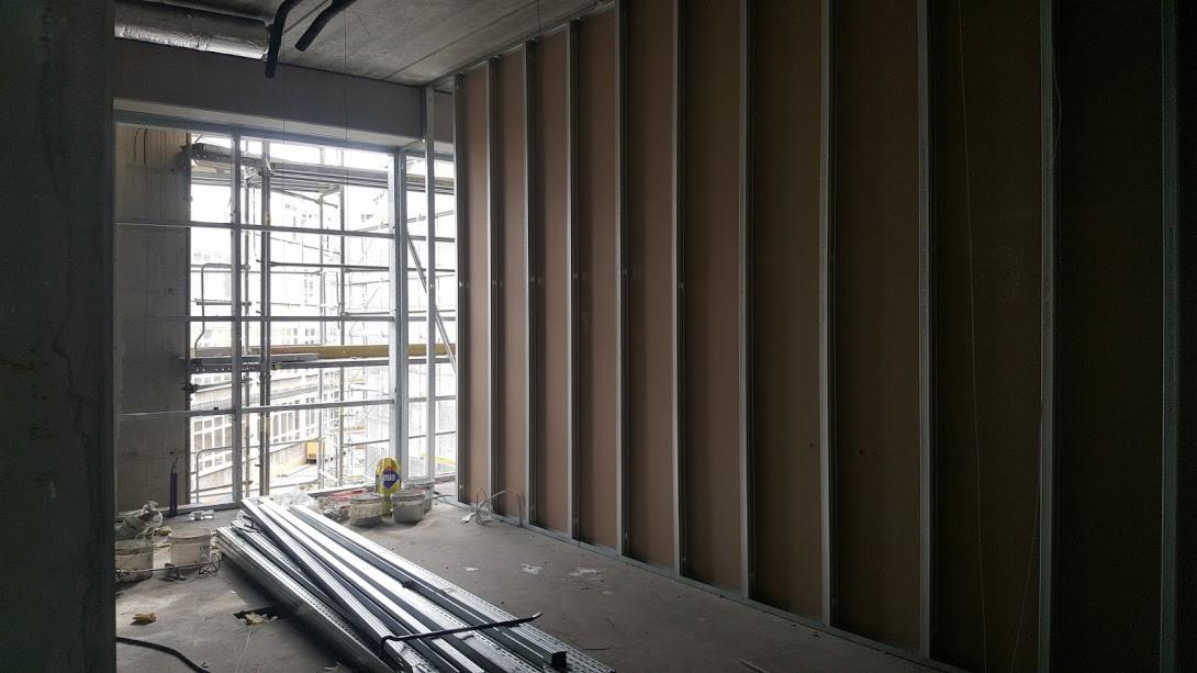 Wnętrze w trakcie szpachlowania ścian z widocznymi profilami i płytami gipsowo-kartonowymi, z oknem na rusztowanie i budynki w tle.
