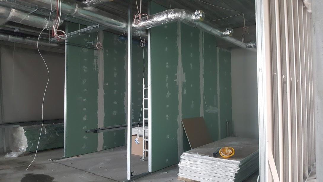 Wnętrze w trakcie montażu ścian działowych z płyt gipsowo-kartonowych, widoczne profile, przewody wentylacyjne pod sufitem oraz stos płyt z żółtym kaskiem ochronnym na wierzchu.