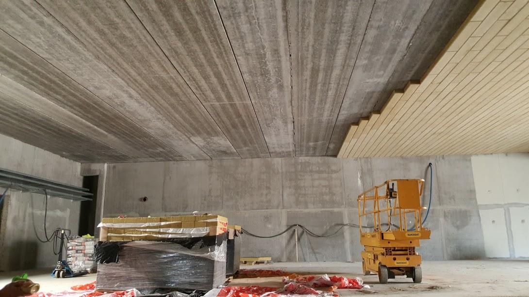 Montaż sufitu: surowy betonowy strop z częściowo zamontowanym jasnym, drewnianym panelem, widoczny podnośnik nożycowy i materiały budowlane na podłodze.