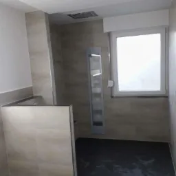 Wykończona łazienka z kabiną prysznicową wyłożoną beżowymi płytkami, ciemna podłoga z widocznymi smugami wody, w tle okno z białymi ramami i wbudowana półka.