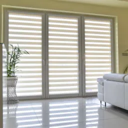 Trzy okna z zamontowanymi beżowo-białymi osłonami okiennymi typu zebra, odbijające się w błyszczącej podłodze, z rośliną doniczkową i fragmentem białej kanapy.