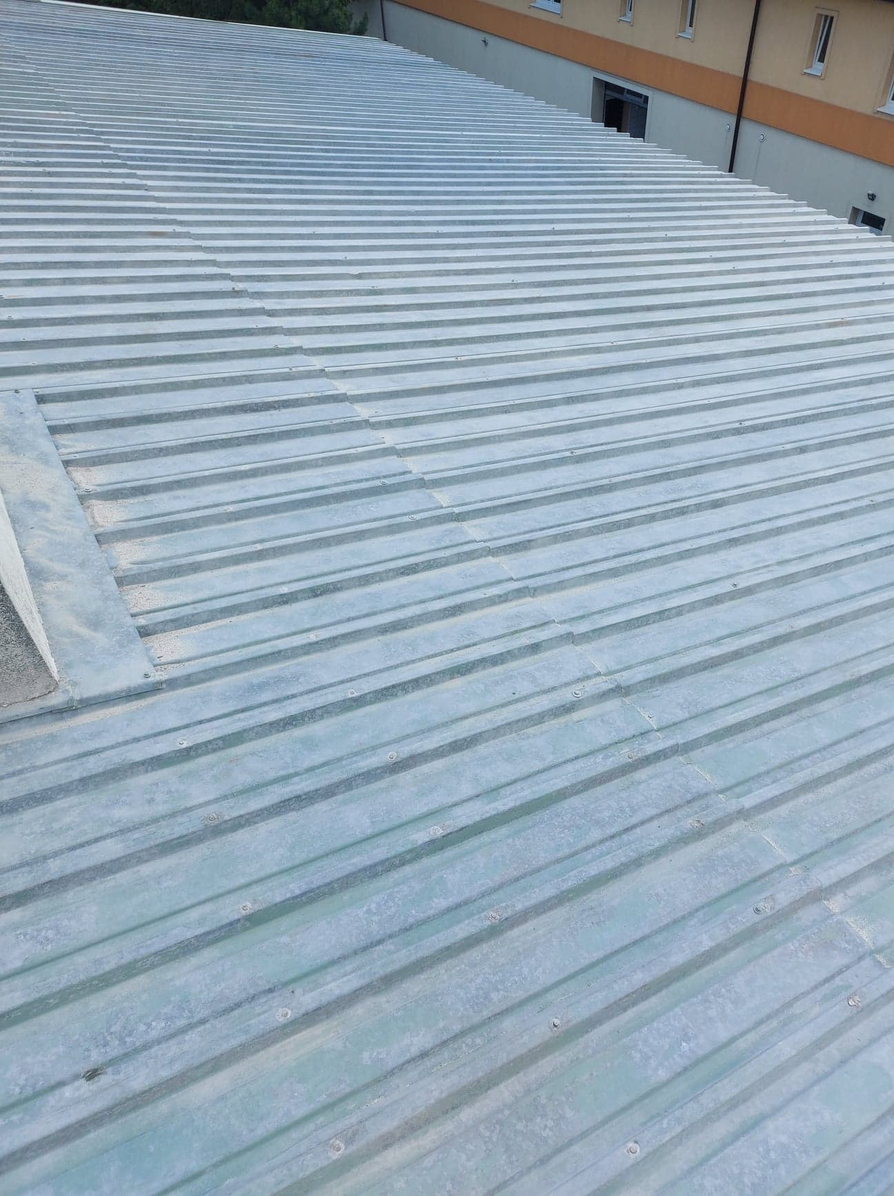 Widok z góry na metalowe pokrycie dachowe z widocznymi śrubami mocującymi i lekkim zabrudzeniem, budynek w tle.