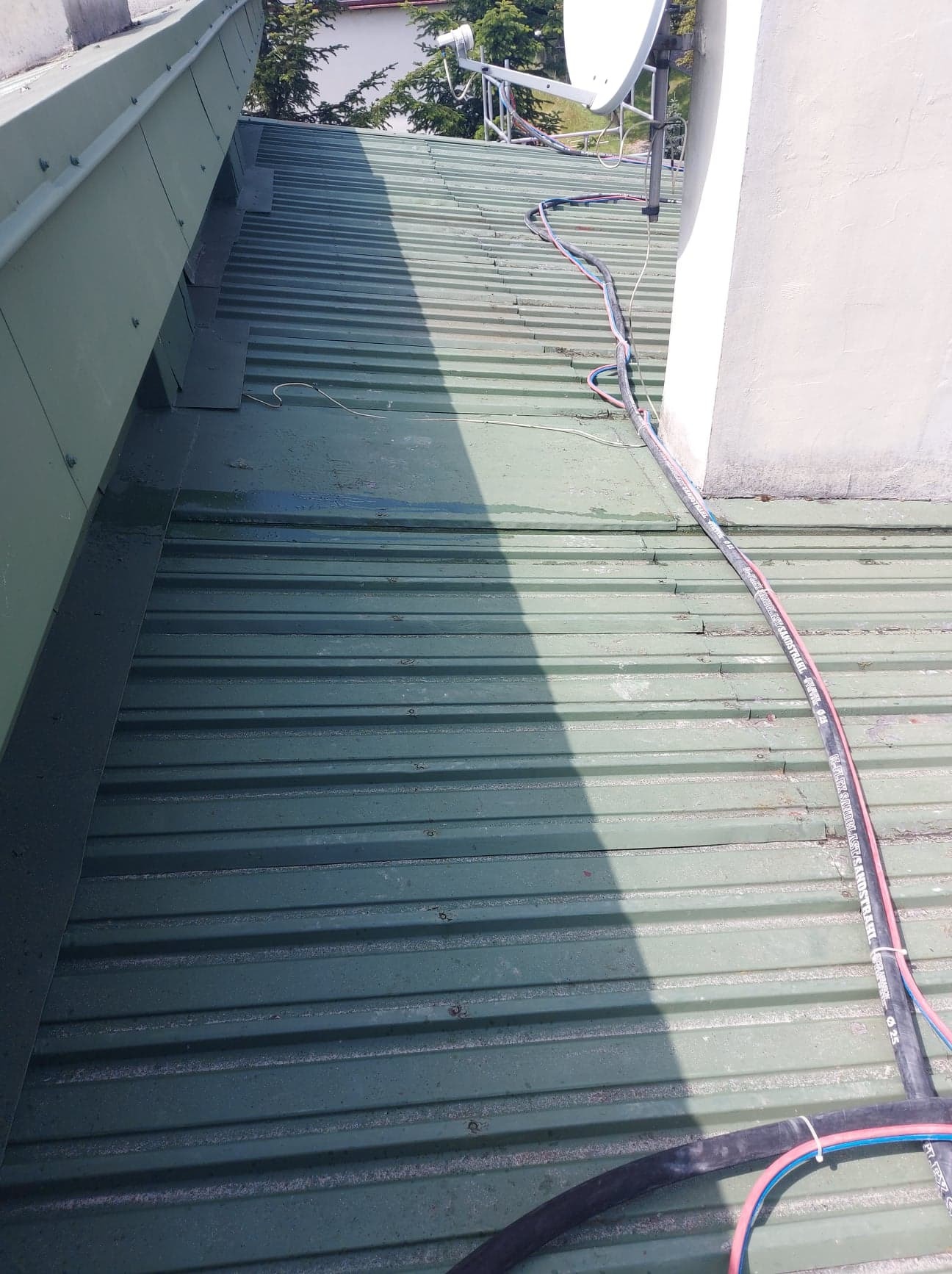 Zielony, blaszany dach z widocznymi przewodami i anteną satelitarną w tle, fragment komina po prawej stronie.