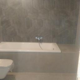 Krzysztof Wojniak - Wykończona łazienka w szarościach: heksagonalne płytki na ścianie nad wanną, bateria wannowa, częściowo widoczna toaleta w folii ochronnej, szare płytki podłogowe.