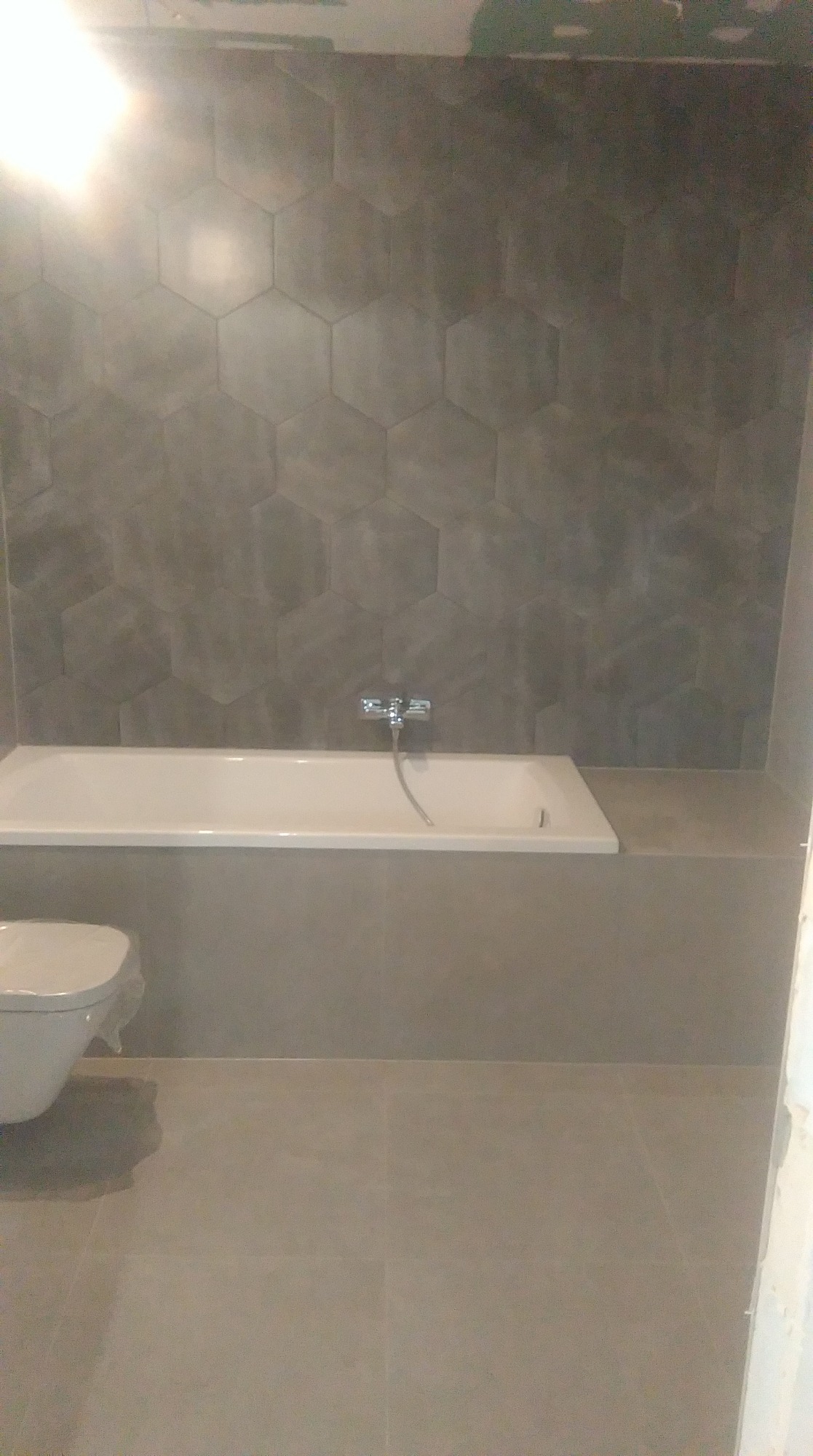 Wykończona łazienka w szarościach: heksagonalne płytki na ścianie nad wanną, bateria wannowa, częściowo widoczna toaleta w folii ochronnej, szare płytki podłogowe.
