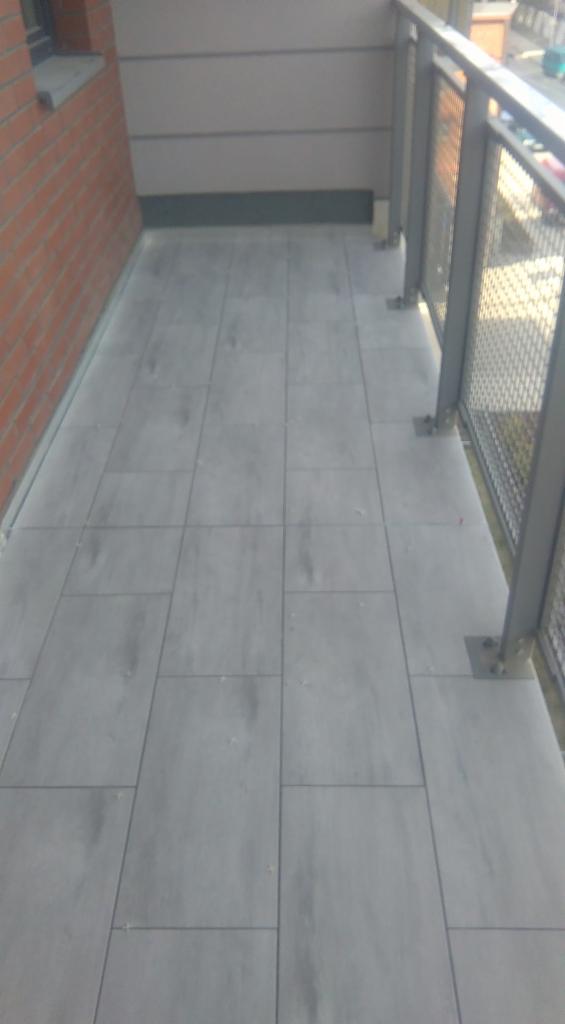 Wyłożony szarymi płytkami balkonik z metalową balustradą i ceglaną ścianą po lewej stronie.