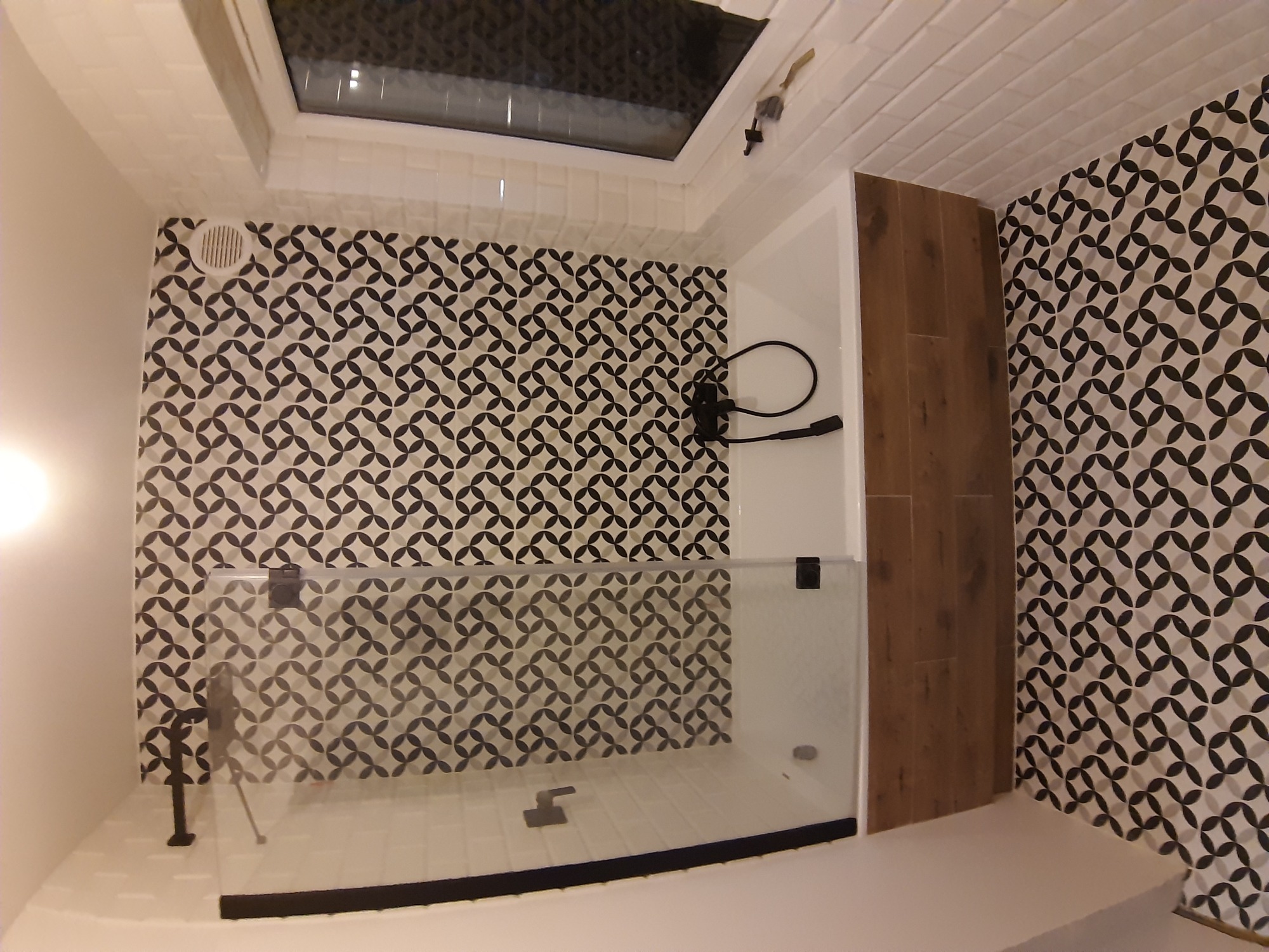 Nowoczesna kabina prysznicowa z czarną armaturą i geometrycznym wzorem płytek na ścianach, oświetlona światłem z okna dachowego.