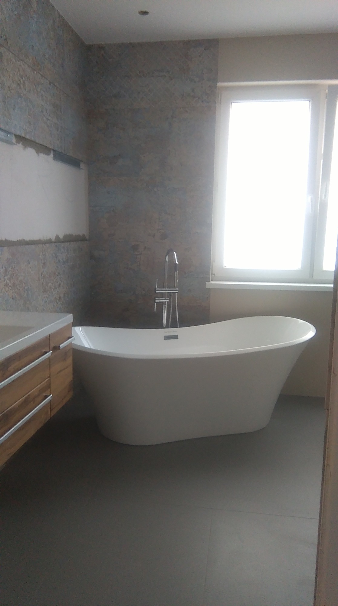 Biała, wolnostojąca wanna z chromowaną armaturą w łazience we Wrocławiu, z fragmentem ściany wykończonej płytkami imitującymi kamień i drewnianą szafką pod umywalkę.