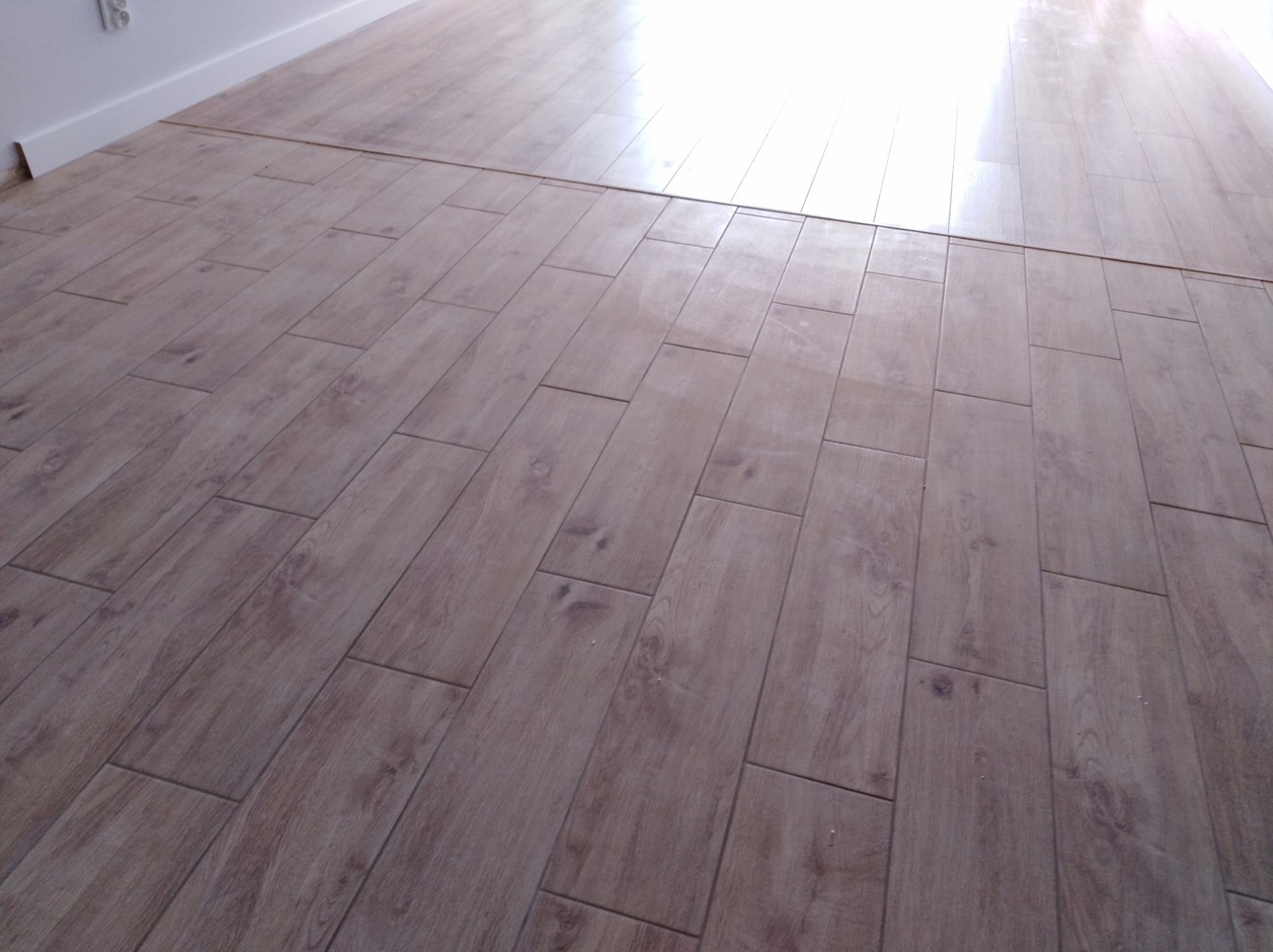 Ułożona podłoga z paneli imitujących drewno, widoczna biała listwa przypodłogowa i fragment podłogi bez paneli, z wyraźnym przejściem między powierzchniami.