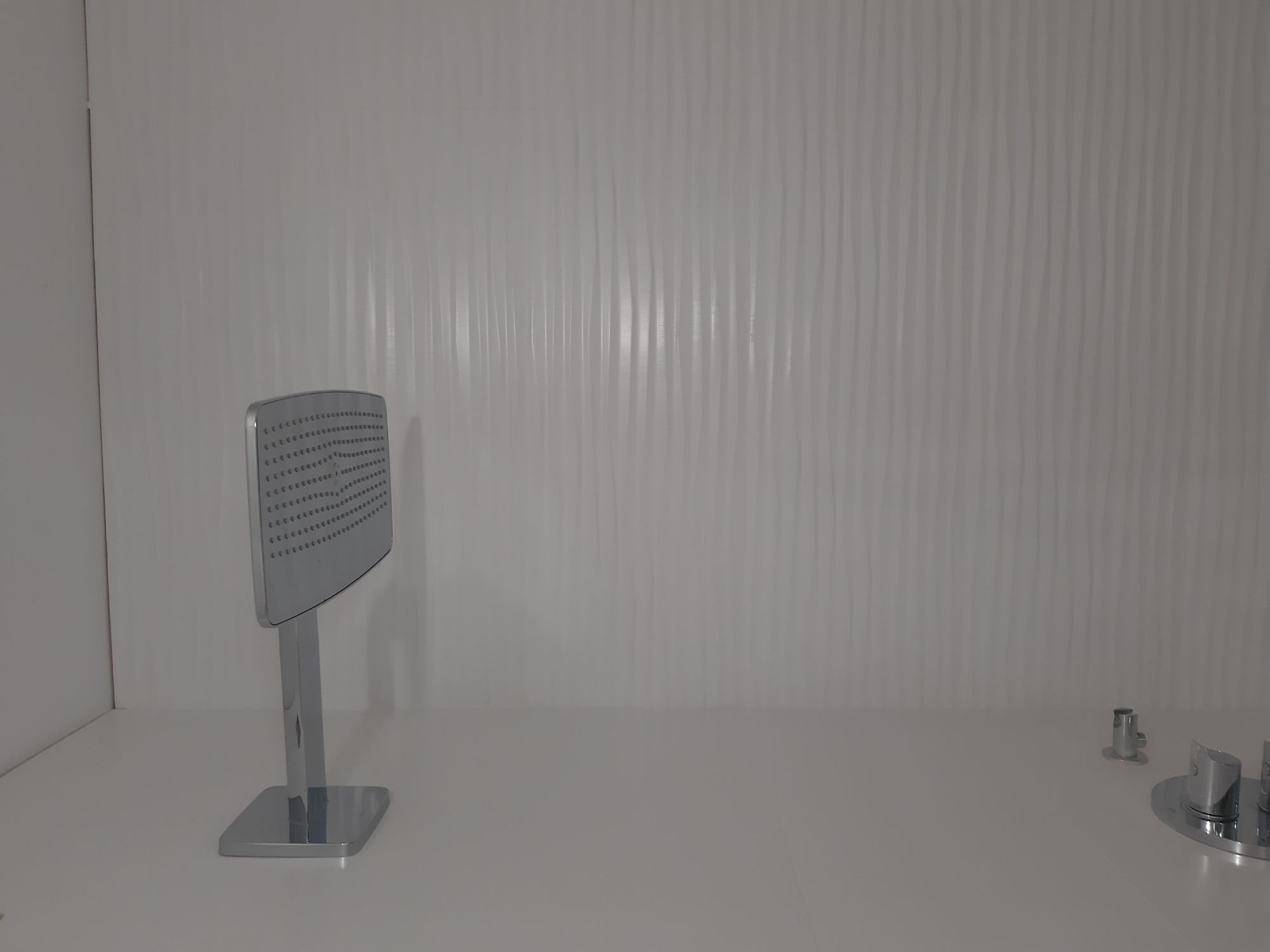 Chromowana deszczownica z kwadratową głowicą i minimalistyczna bateria umywalkowa na tle białej ściany z falistą strukturą.