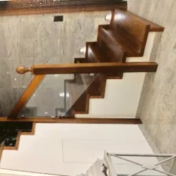 Drewniane schody z białymi podstopnicami i szklaną balustradą, widok z góry, na tle marmurowej ściany.