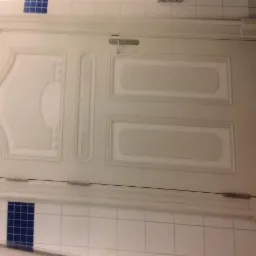 Białe, wewnętrzne drzwi z dekoracyjnymi panelami, zamontowane w łazience z biało-niebieskimi płytkami, widoczne zawiasy i klamka.