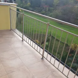 Wykończony balkon z beżowymi płytkami i metalową balustradą, widok na zielony ogród.