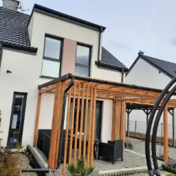 Nowoczesny dom z drewnianą pergolą i meblami ogrodowymi na brukowanym tarasie, widok z boku na tle pochmurnego nieba.