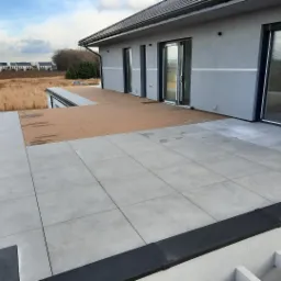 Nowoczesny taras z szarych płyt betonowych i drewnopodobnej deski kompozytowej, widok z góry na częściowo ukończoną konstrukcję, w tle zabudowa jednorodzinna i łąka.