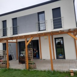 BRADERS Jarosław Słomiński - Nowoczesny dom z szarą elewacją i drewnianą pergolą z jasnego drewna, widoczne dzieci w drzwiach tarasowych, drabina oparta o ścianę, zabawki na tarasie.