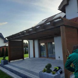 BRADERS Jarosław Słomiński - Zadaszony taras z drewnianą konstrukcją, wykończony szarą kostką brukową, z widocznymi elementami domu i ogrodu. Osoba w zielonej bluzie maluje drewniane elementy konstrukcji.