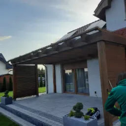 Zadaszony taras z drewnianą konstrukcją, wykończony szarą kostką brukową, z widocznymi elementami domu i ogrodu. Osoba w zielonej bluzie maluje drewniane elementy konstrukcji.