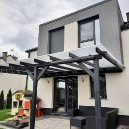 BRADERS Jarosław Słomiński - Nowoczesna pergola z ciemnego drewna z mlecznym zadaszeniem przylegająca do jasnego domu z szarymi elementami architektonicznymi, na tarasie czarne meble ogrodowe, obok domek dla dzieci.