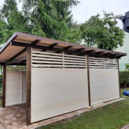 Drewniana konstrukcja z brązowym dachem, jasnymi deskami i poziomymi szczelinami, stojąca na kostce brukowej obok trawnika.