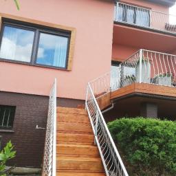 BRADERS Jarosław Słomiński - Schody zewnętrzne z drewna prowadzące na balkon z metalową balustradą, widok na dwupiętrowy budynek z oknami w drewnianych ramach i roślinnością.