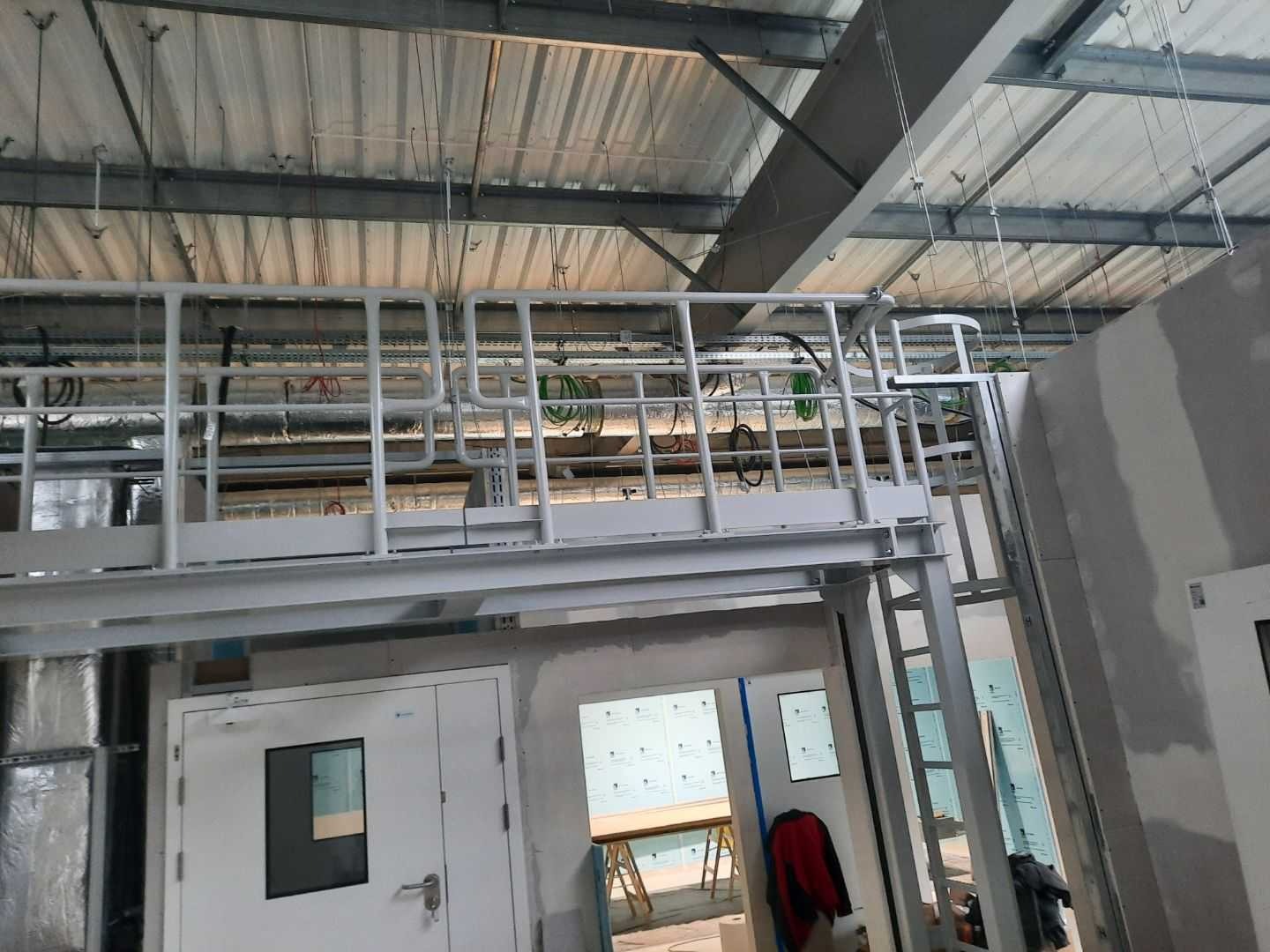 Wnętrze w trakcie prac wykończeniowych: metalowa konstrukcja antresoli z balustradą, widoczne instalacje pod sufitem, surowe ściany i drzwi z szybą.