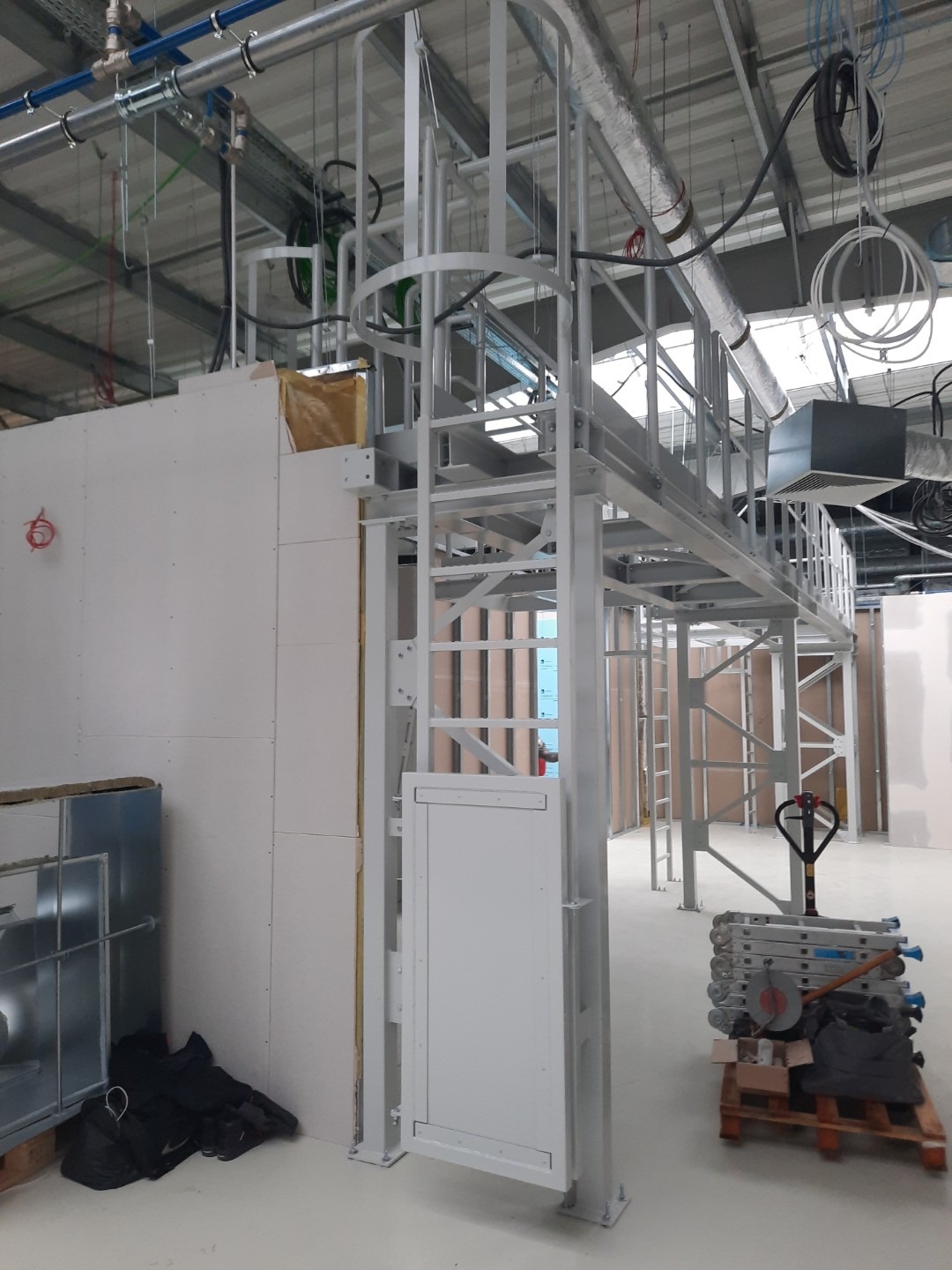 Wnętrze budynku w trakcie remontu: widoczna stalowa konstrukcja pomostu roboczego z drabinką i klapą, ściany z płyt gipsowo-kartonowych, instalacje pod sufitem i narzędzia na palecie.