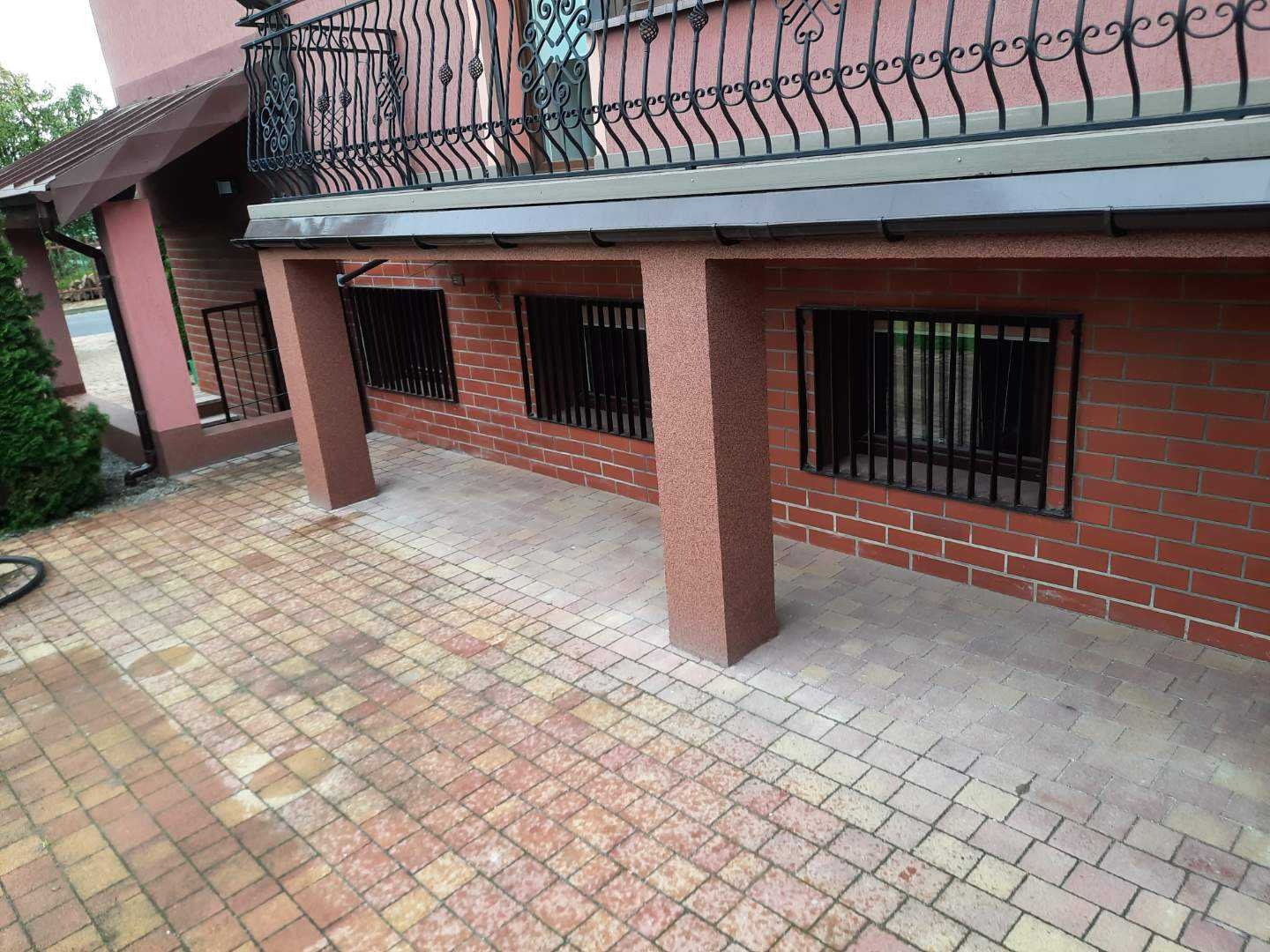 Ceglana podmurówka budynku z kratami w oknach piwnicznych i wyłożony kostką brukową taras.