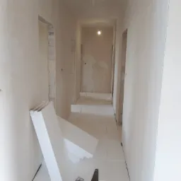 Korytarz w trakcie prac wykończeniowych, ściany przygotowane pod malowanie, na podłodze i przy ścianie leżą płyty styropianowe, widoczne kable elektryczne wystające ze ściany.