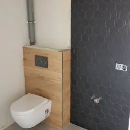 Podwieszana toaleta z drewnianą obudową stelaża, czarne heksagonalne płytki na ścianie, szare płytki podłogowe, widoczne elementy instalacji hydraulicznej.