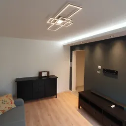 Nowoczesny salon po remoncie: szara sofa z poduszką, czarne komody, drewniane panele podłogowe, ciemnoszara ściana z zamontowanym uchwytem na telewizor i listwą LED pod sufitem, geometryczna lampa...