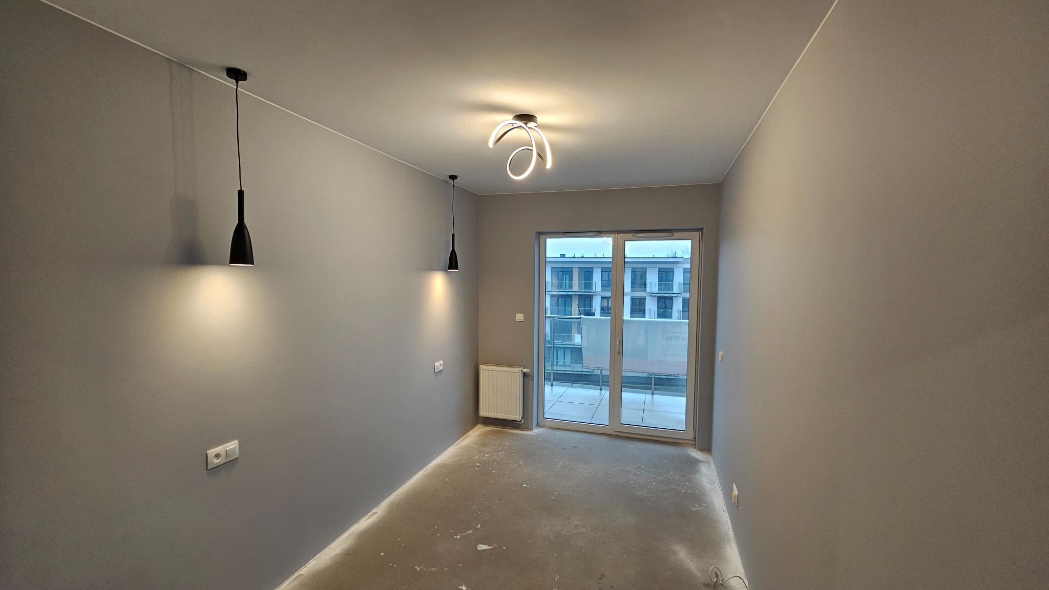 Surowe wnętrze szarego pokoju z zamontowanymi lampami wiszącymi i nowoczesną lampą sufitową, widoczne gniazdka elektryczne i grzejnik, przygotowane do dalszych prac wykończeniowych.