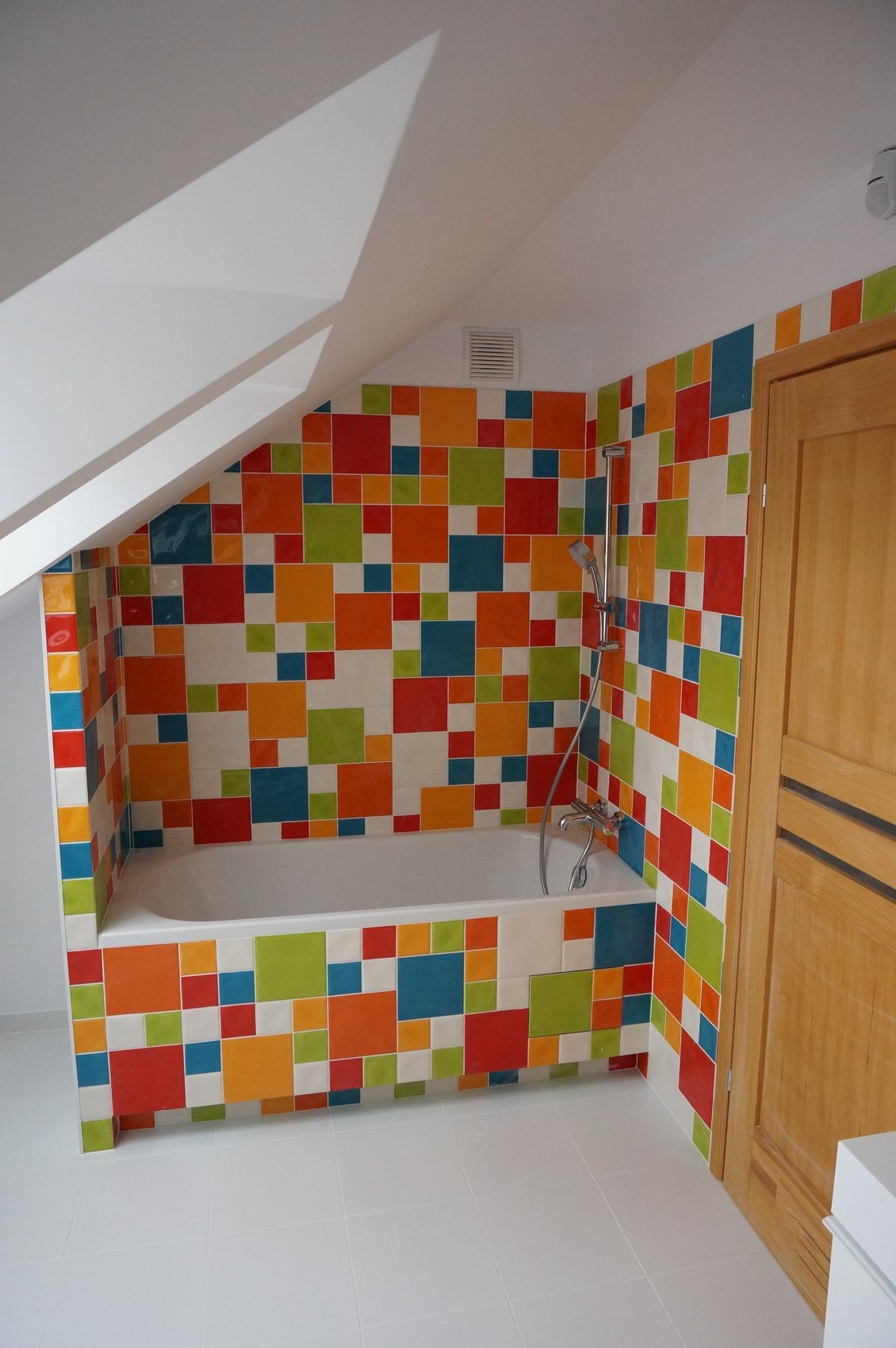 Wykończona łazienka na poddaszu z wanną obudowaną kolorowymi, kwadratowymi płytkami, armatura prysznicowa zamontowana na ścianie, biała podłoga i drewniane drzwi.