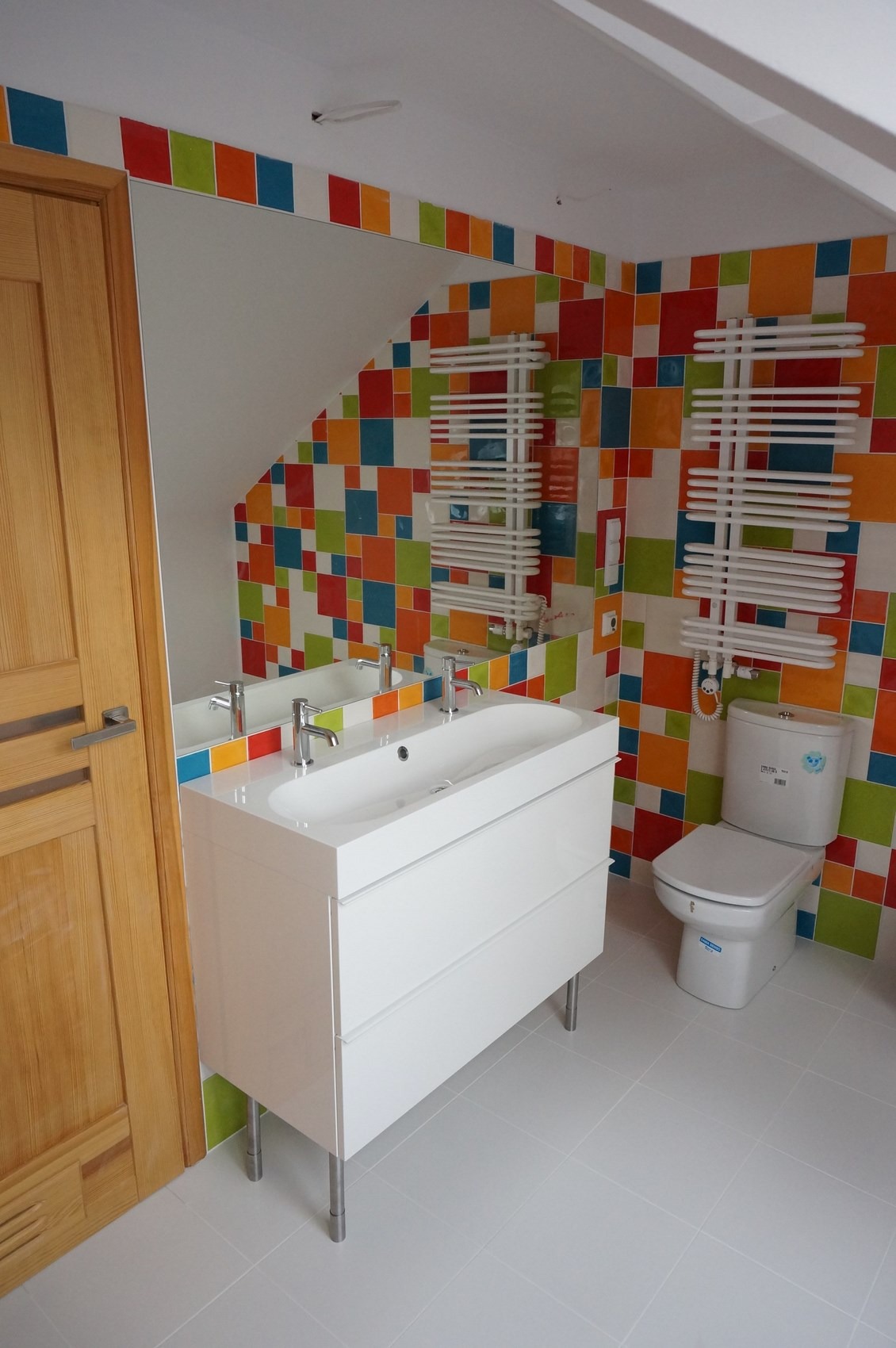 Jasna łazienka z białą szafką umywalkową z dwiema bateriami, toaletą, grzejnikami drabinkowymi i ścianami wyłożonymi kolorowymi, kwadratowymi płytkami.