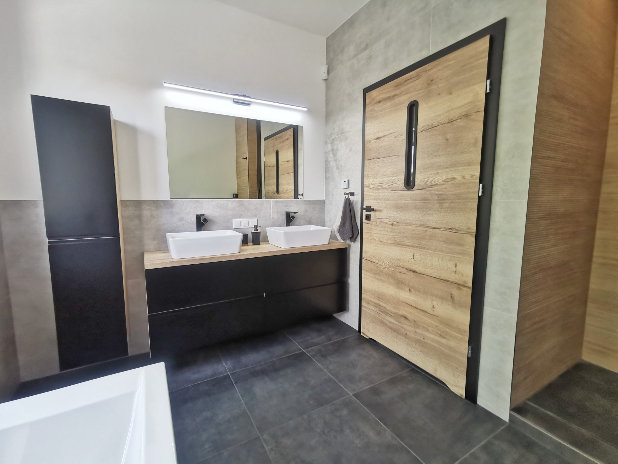Nowoczesna łazienka z dwoma białymi umywalkami nablatowymi, czarną szafką pod umywalką, drewnianymi drzwiami z czarną ramą i podłużnym okienkiem, szarymi płytkami na ścianach i podłodze...