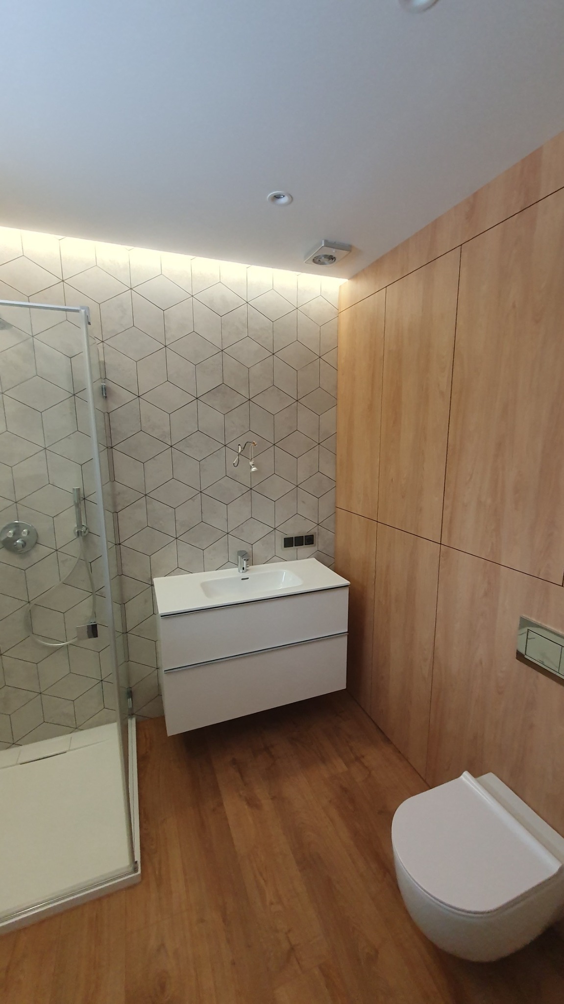 Nowoczesna łazienka z kabiną prysznicową, białą szafką z umywalką, toaletą i drewnianą zabudową ścienną. Ściana wyłożona płytkami w geometryczny wzór 3D.