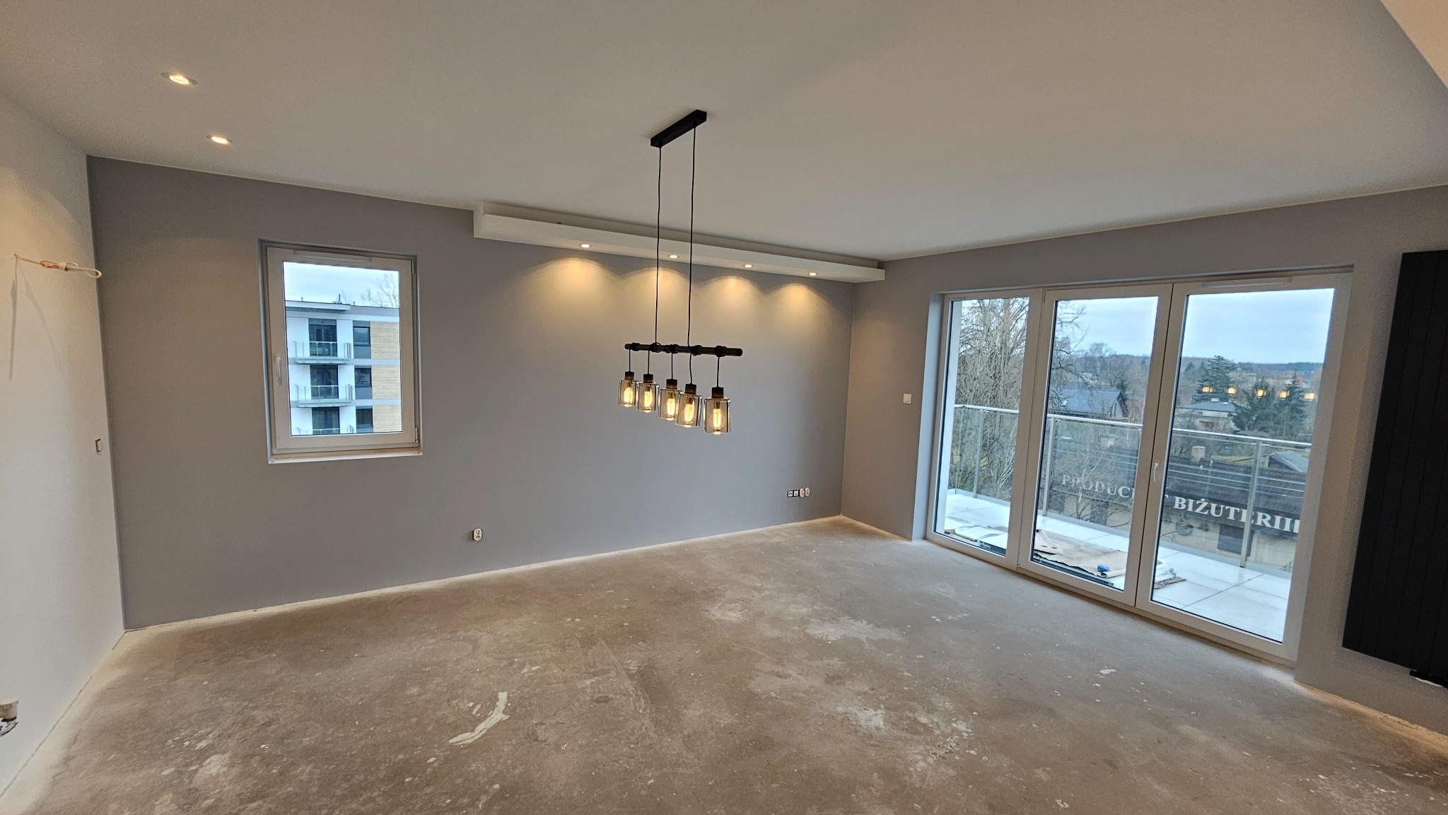 Pokój w trakcie wykańczania: szare ściany, surowa podłoga, nowoczesna lampa wisząca z pięcioma szklanymi kloszami, okno z widokiem na budynek i balkon z balustradą, duże okno balkonowe.