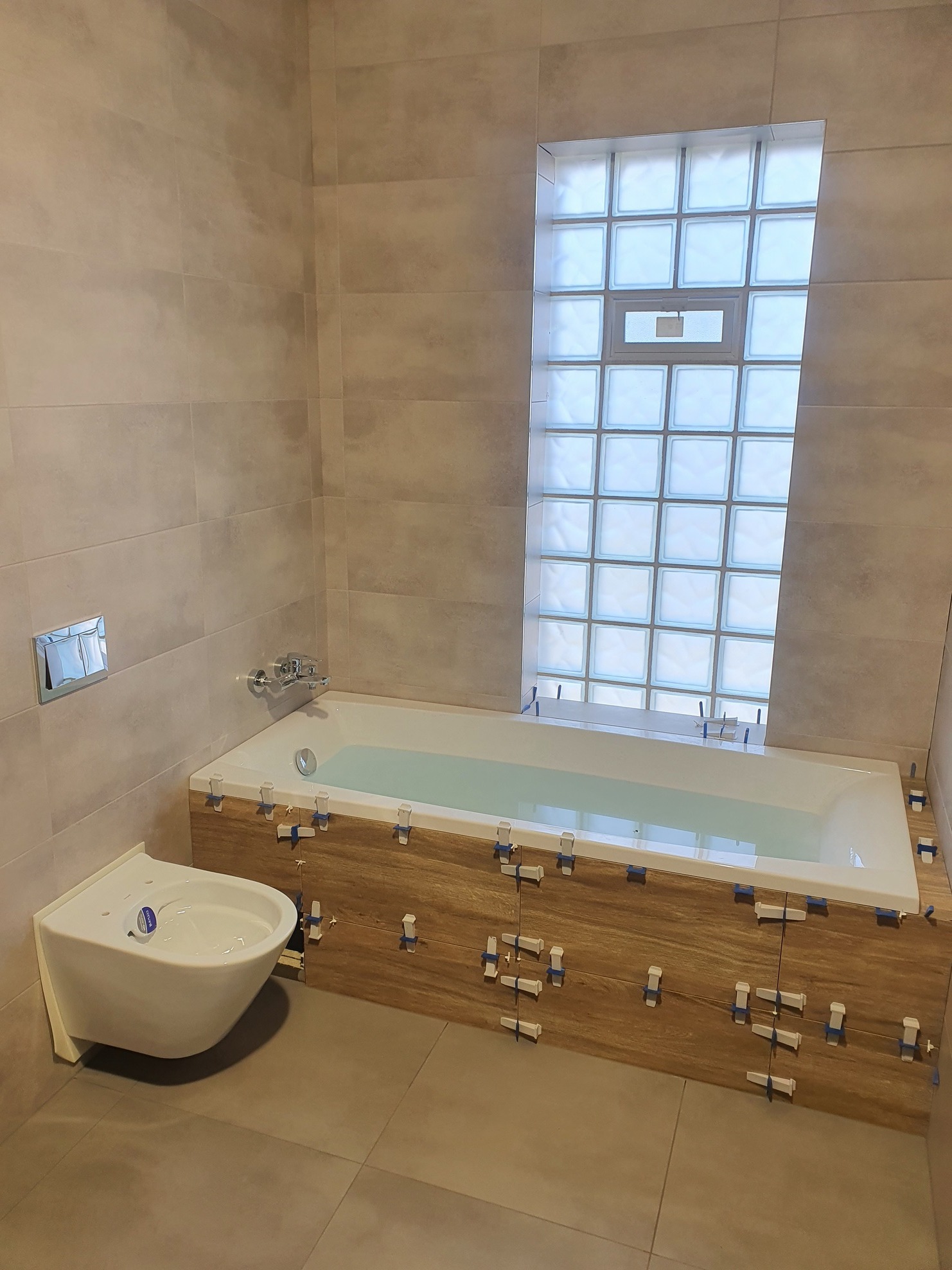 Biała wanna wypełniona wodą, obudowana panelami imitującymi drewno, z widocznymi krzyżykami dystansowymi, obok podwieszana toaleta, ściany wyłożone płytkami w odcieniach beżu, okno z luksferów.