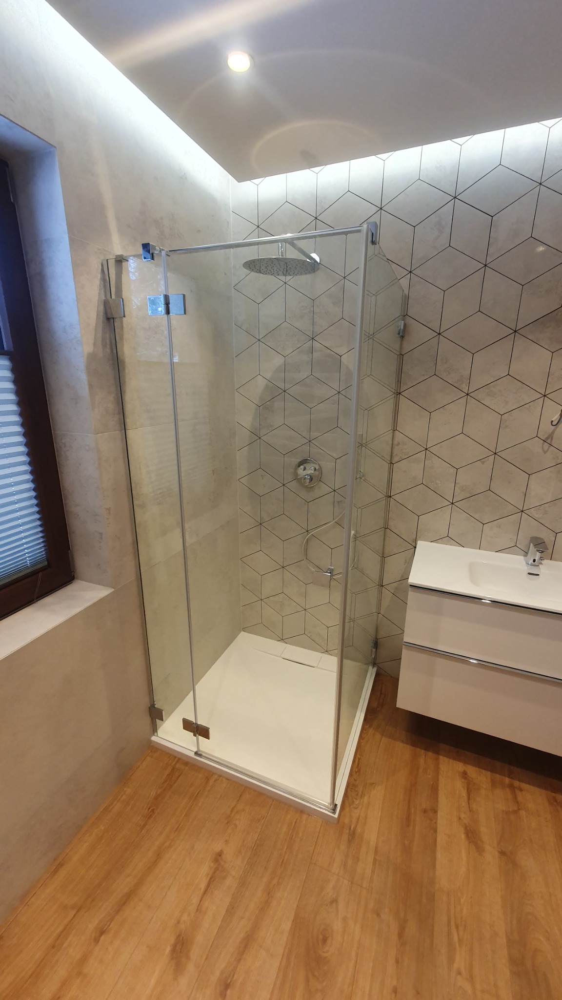 Nowoczesna kabina prysznicowa z deszczownicą w łazience wykończonej płytkami w geometryczne wzory i drewnopodobną podłogą, obok umywalka z szafką.