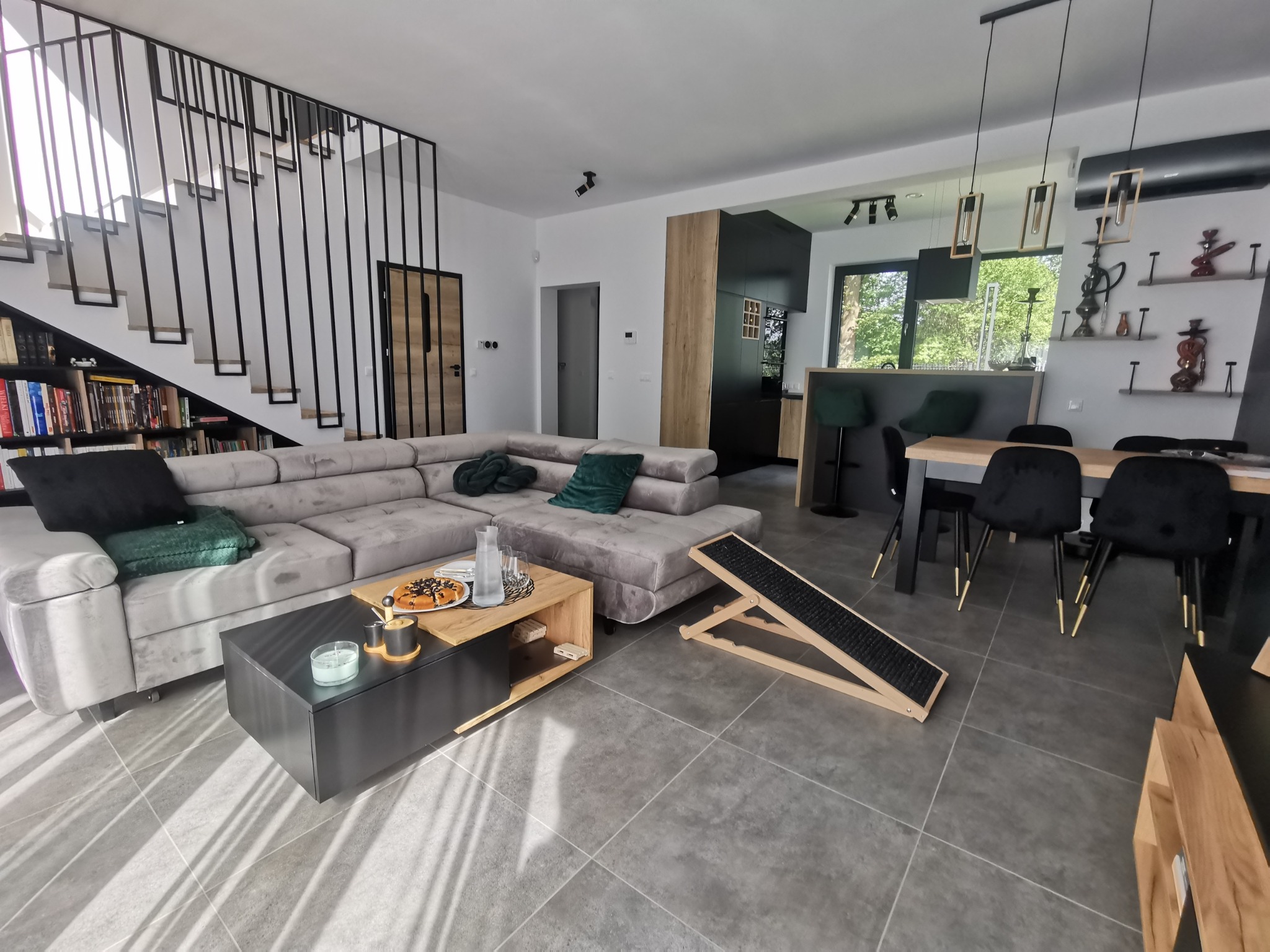 Przestronny salon z szarą sofą narożną, czarnym stolikiem kawowym z dekoracjami, drewnianymi schodami z czarną balustradą oraz otwartą kuchnią z jadalnią w tle. Widoczny również drewniany podjazd...