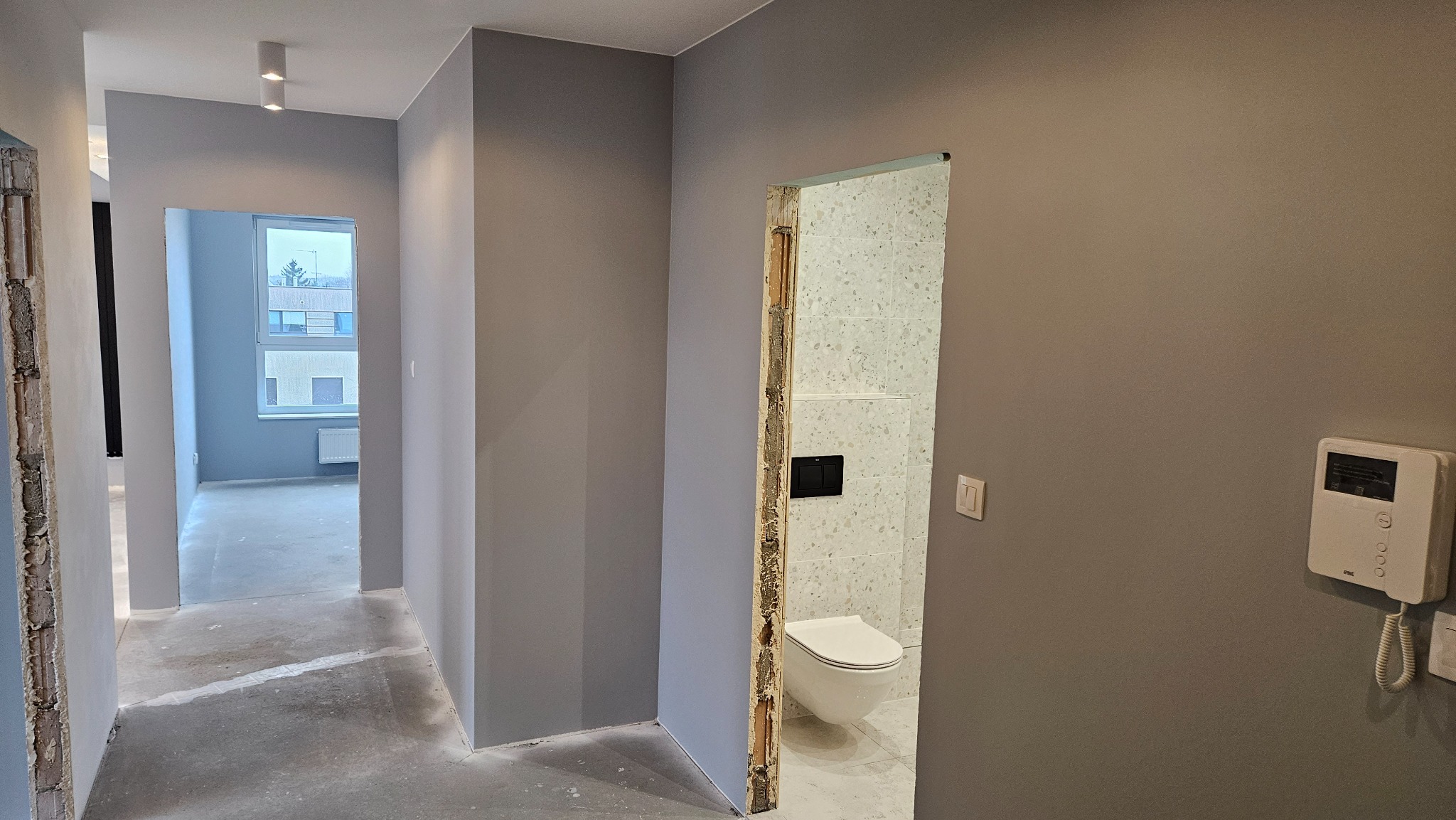 Wnętrze mieszkania w trakcie prac wykończeniowych: szare ściany, surowa podłoga, widok na łazienkę z toaletą przez niedokończone futryny, w tle widok na pokój z oknem.
