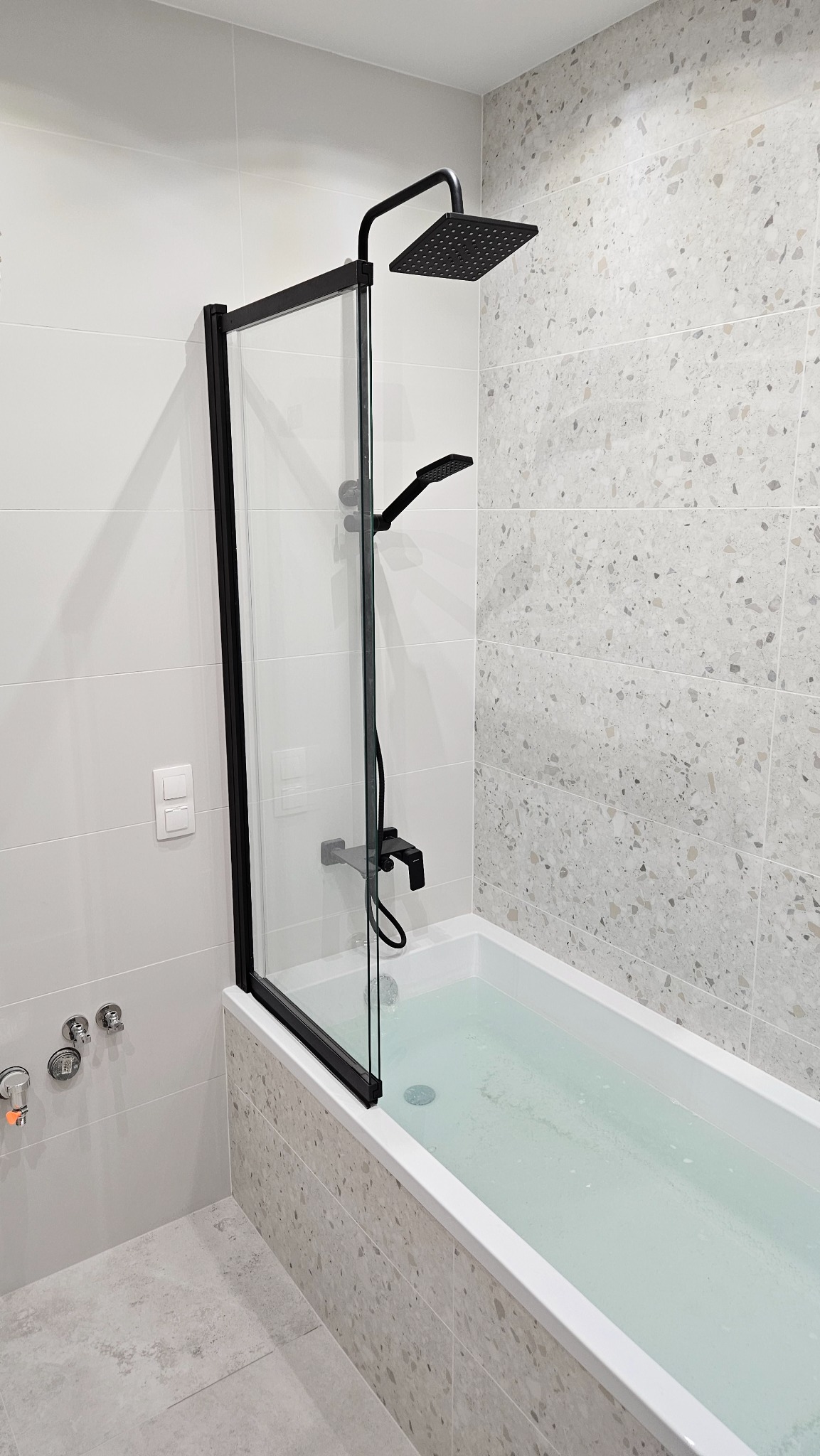 Wanna wypełniona wodą z zamontowaną czarną armaturą i szklanym parawanem, ściany wyłożone płytkami w odcieniach szarości i bieli.