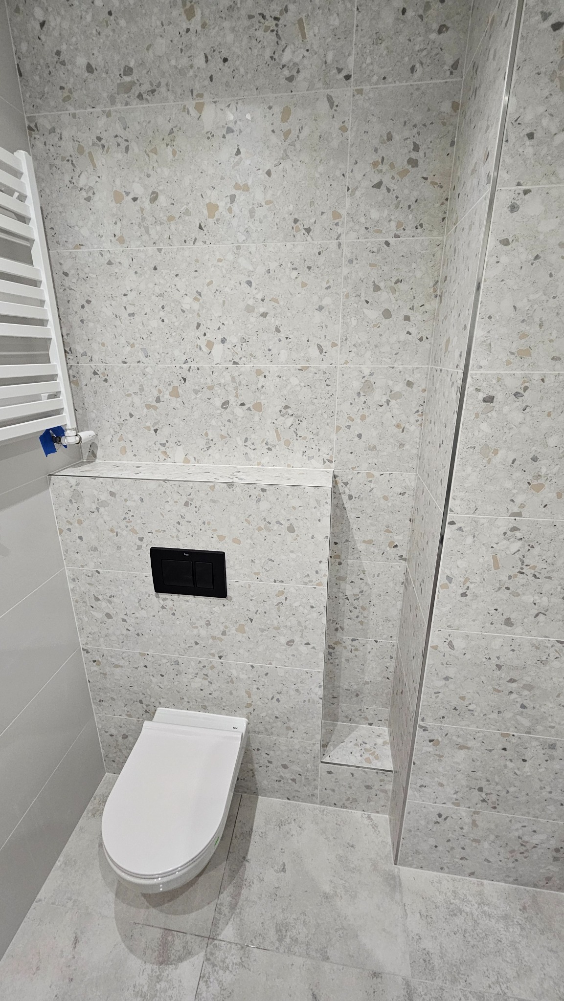 Nowoczesna toaleta podwieszana z czarnym przyciskiem spłukującym na tle ściany wyłożonej płytkami imitującymi lastryko, widoczny grzejnik drabinkowy po lewej stronie.