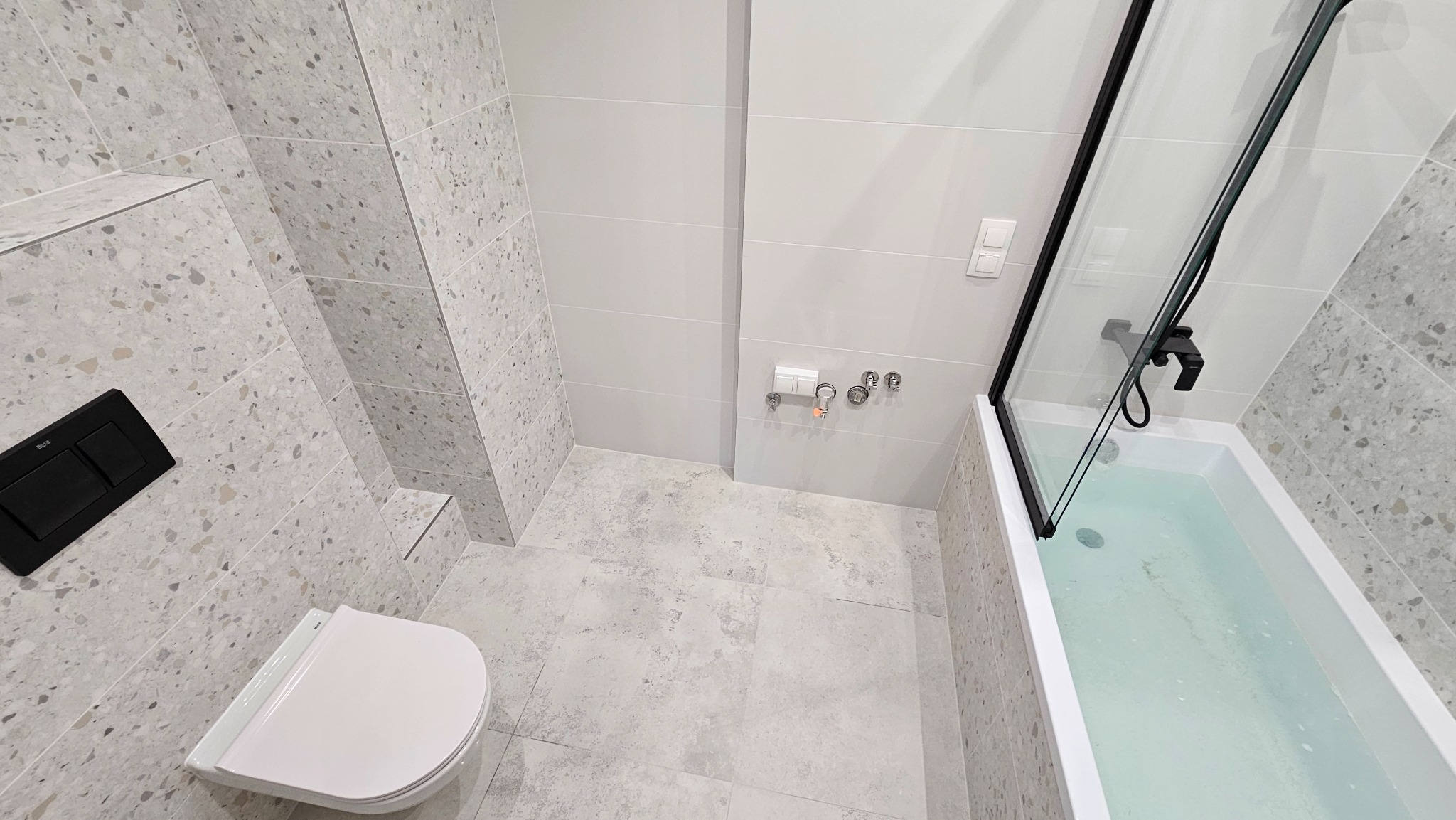 Nowoczesna łazienka w jasnych odcieniach szarości z terazzo na ścianach, z wiszącą toaletą z czarnym przyciskiem spłukiwania i wanną wypełnioną wodą z czarną armaturą oraz szklanym parawanem.