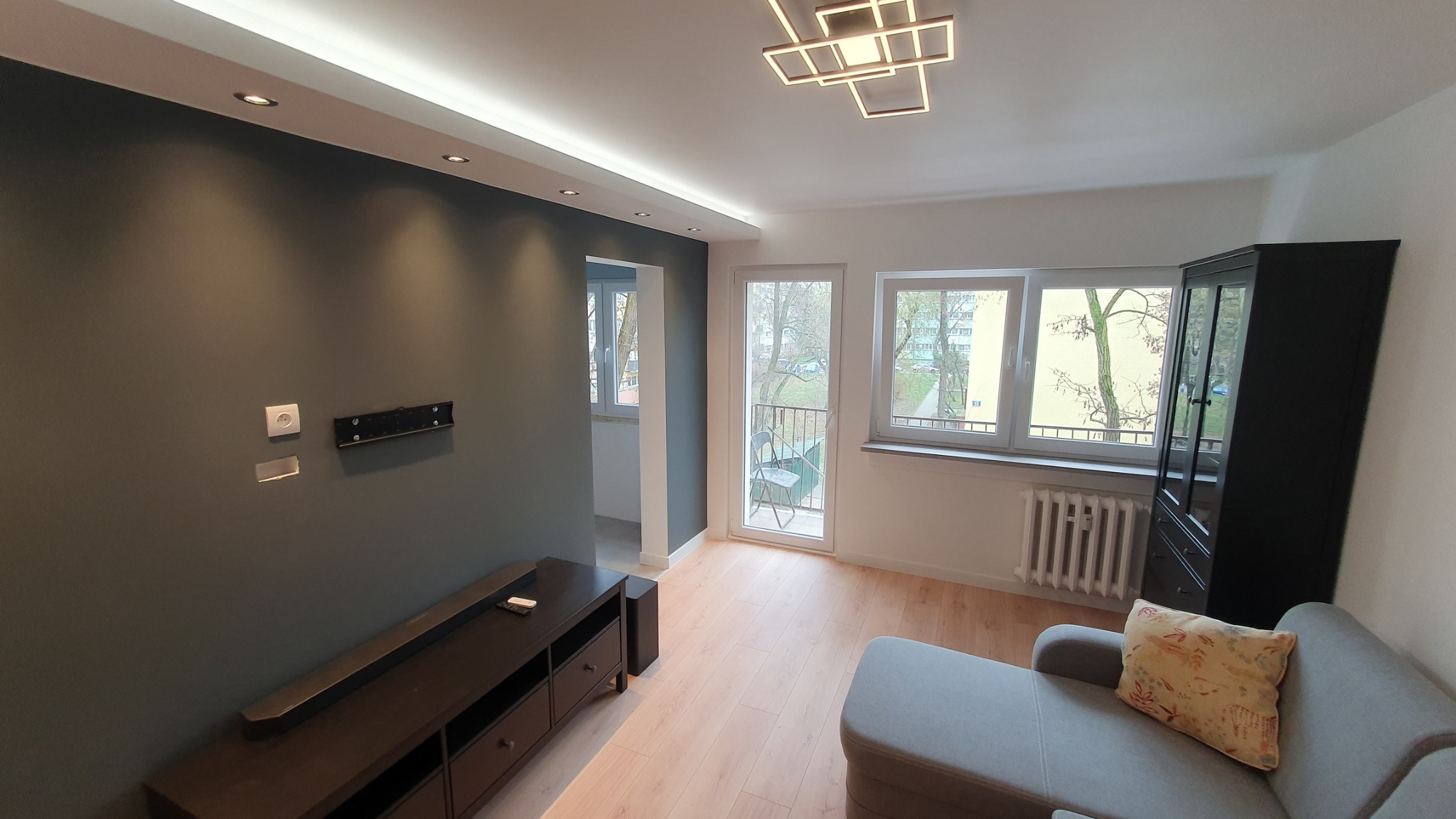 Jasne wnętrze pokoju po remoncie: szara ściana z gniazdkiem i uchwytem na telewizor, podłoga z paneli, nowoczesna lampa sufitowa, okno balkonowe z widokiem na zewnątrz.