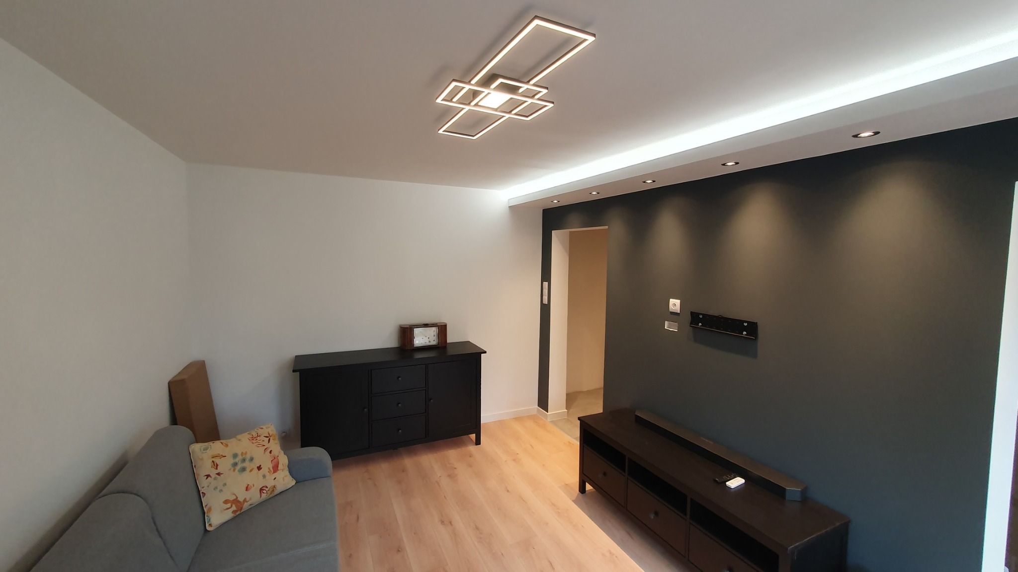 Nowoczesny salon po remoncie: szara sofa z poduszką, czarne komody, drewniane panele podłogowe, ciemnoszara ściana z zamontowanym uchwytem na telewizor i listwą LED pod sufitem, geometryczna lampa...