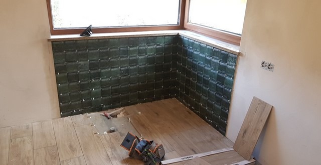 Proces układania ciemnozielonych, kwadratowych płytek na ścianie w rogu pomieszczenia, z widocznymi krzyżykami dystansowymi i narzędziami glazurniczymi na podłodze z paneli.