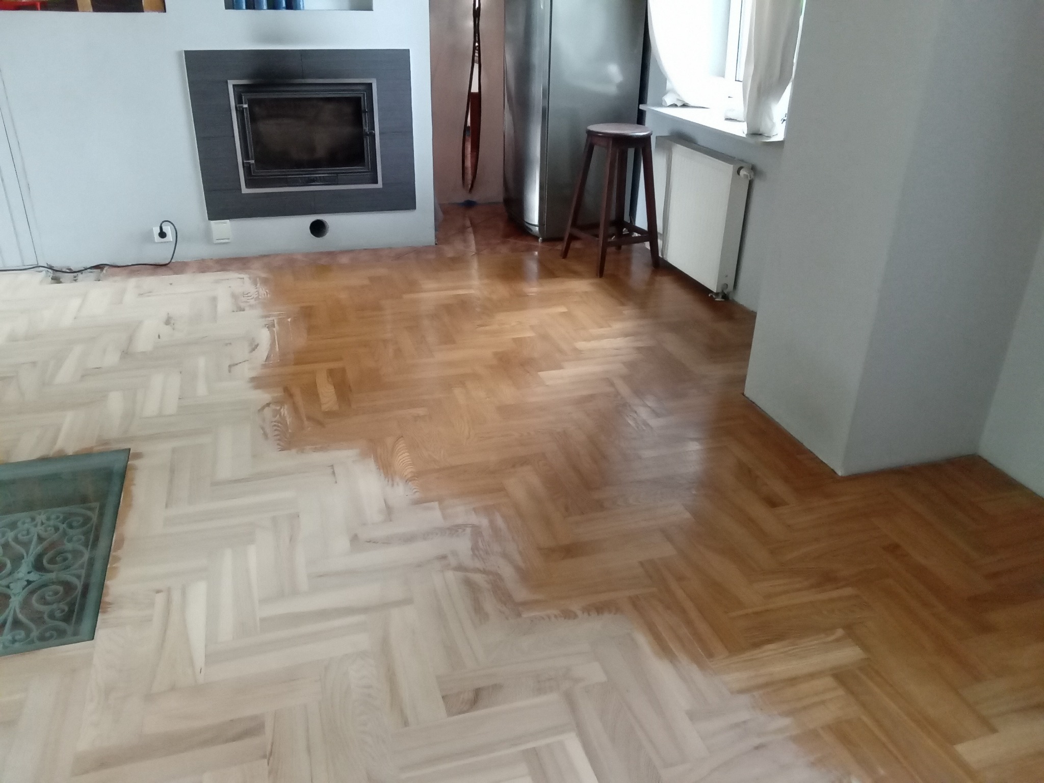 Częściowo wycyklinowana podłoga parkietowa w jodełkę, widoczna różnica w kolorze między surowym a odnowionym drewnem, fragment wnętrza z kominkiem i stołkiem w tle.