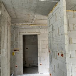 PS Energia Piotr Sitnik - Wnętrze w stanie surowym z widocznymi przewodami elektrycznymi poprowadzonymi po ścianach z betonu komórkowego oraz pod sufitem, zabezpieczonymi pianką montażową w narożnikach.